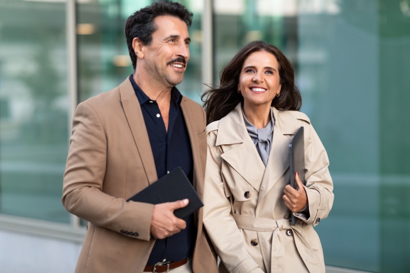 Cheerful business partners walking by street, going to business meeting. Middle aged man and woman wearing formal outfits, holding gadgets in their hands, coming back to office